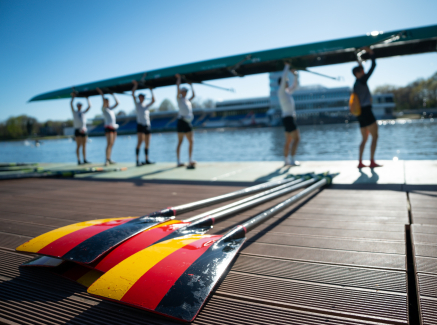 Ruderer mit dem Boot über dem Kopf, im Vordergrund Ruderblätter in den Deutschland-Farben