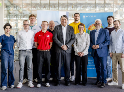 Elf Personen in einem Halbkreis vor zwei Bannern, hinter ihnen eine Trennwand aus Glasbausteinen