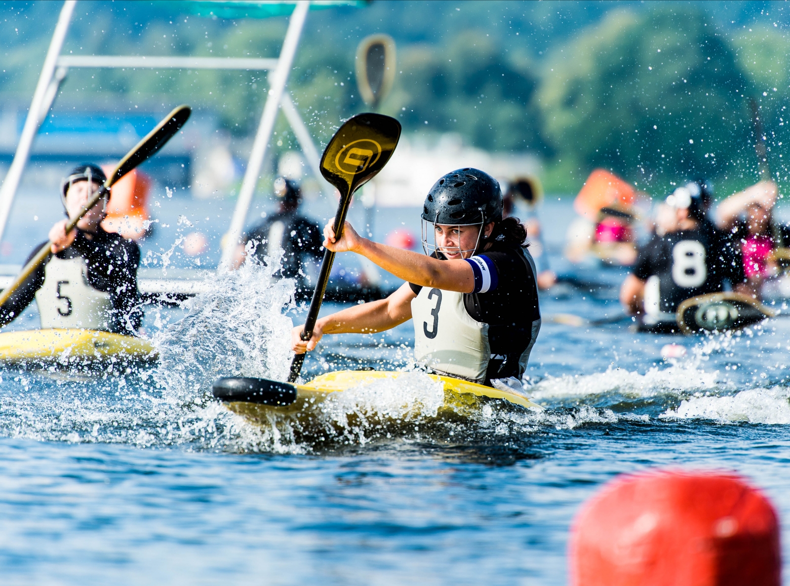 Frauen beim Kanupolo. Eine Frau mit einem schwazren Helm und schwarz-weißen Trikot mit der Nummer 3 prescht paddelnd mit ihrem gelb-schwarzen Kanu vorwärts, das Wasser spritzt nach allen Seiten.