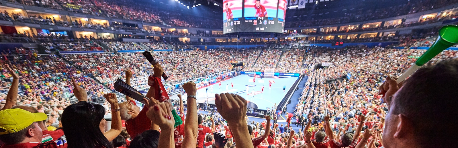 Man sieht von der hinteren Reihe in dievollbesetzte, geschlossene Mehrzweckhalle Lanxess Arena. Viele Zuschauer haben den rechten Arm oben und jubeln ihrer Mannschaft zu. Oben in der Mitte hängt eine kreisrunde Bildschirmpräsentator an der Decke.