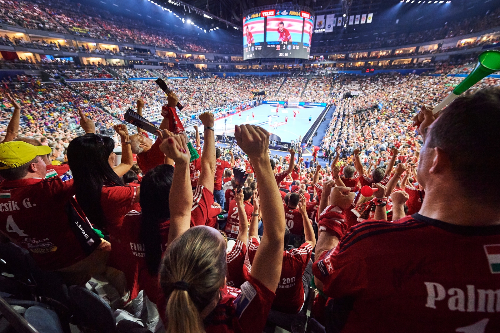 Man sieht von der hinteren Reihe in dievollbesetzte, geschlossene Mehrzweckhalle Lanxess Arena. Viele Zuschauer haben den rechten Arm oben und jubeln ihrer Mannschaft zu. Oben in der Mitte hängt eine kreisrunde Bildschirmpräsentator an der Decke.