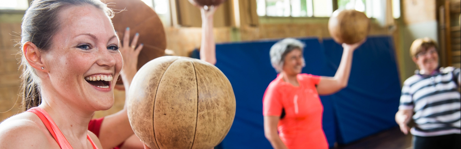 Lächelnde Frau in rosa-pinker Sportbekleidung hält einen braunen Meidzinball hoch. Im Hintergrund stehen noch weitere Sportlerinnen mit Medizinbällen.