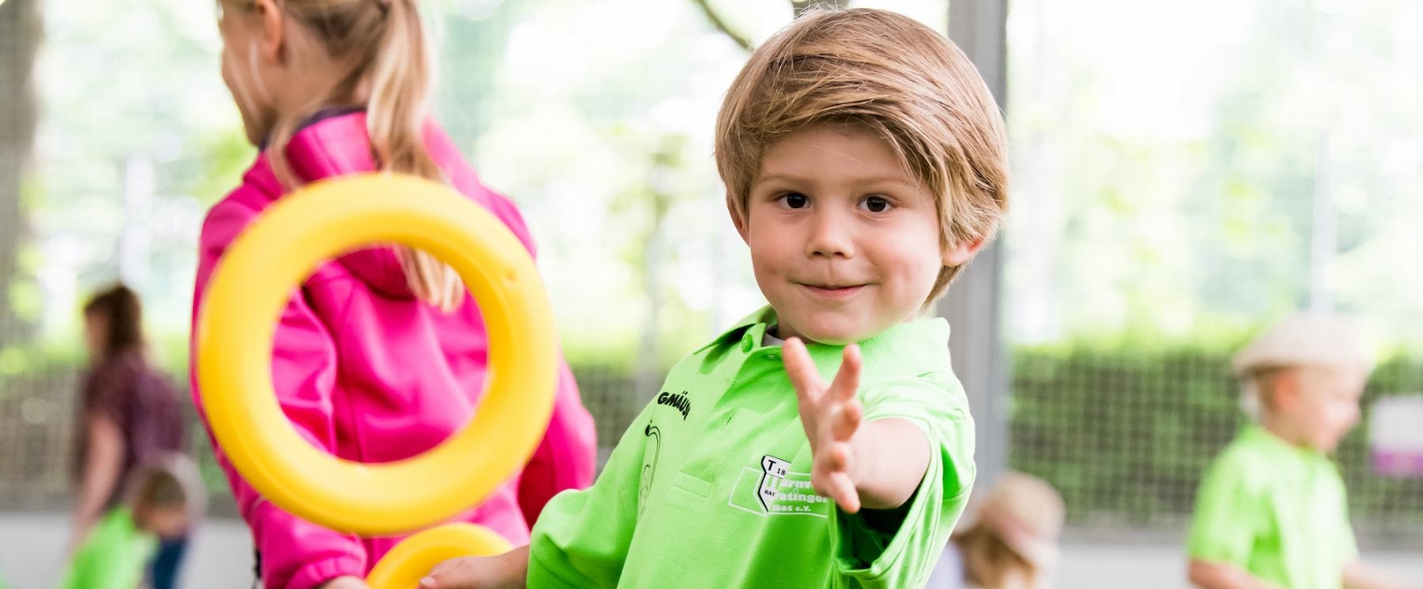 Ein kleiner, blonder Junge im grünen Hemd streckt lächelnd seine linke Hand nach der Kamera aus.