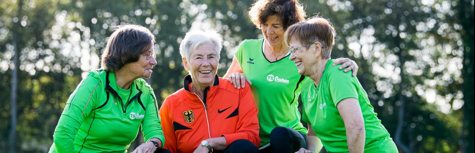 Vier ältere Damen bei einer Teambesprechung beim Sport. Drei Damen haben ein grünes Oberteil an, eine Dame in der Mitte hat ein rotes Oberteil an. Alle haben schwarze Sporthosen an.