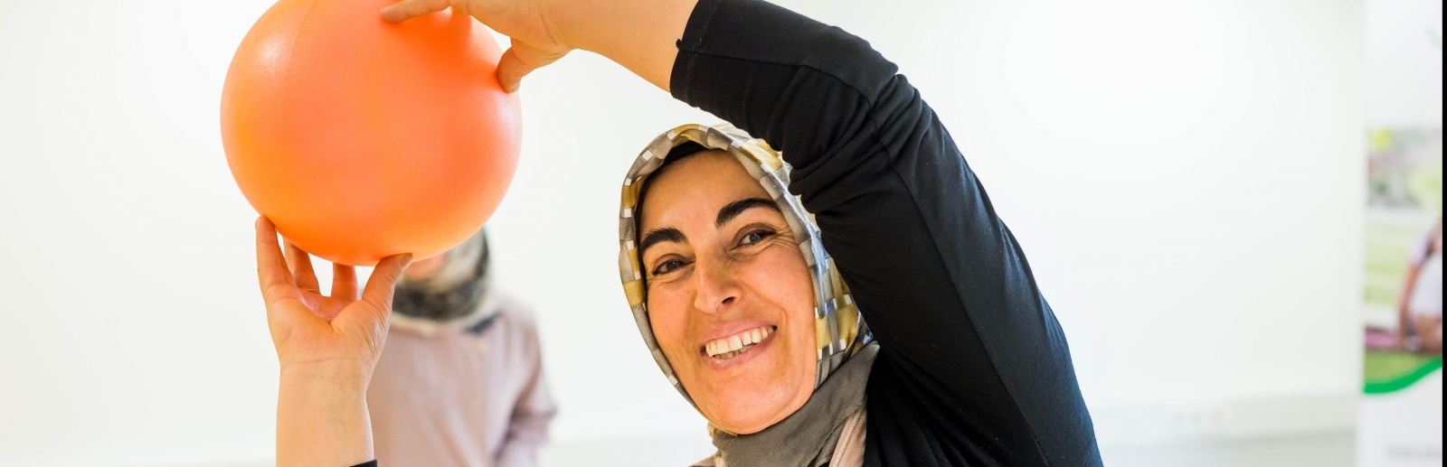 Eine lächelnde Frau mit schwarzem Oberteil und beigefarbenen gemusterten Kopftuch hält bei einer Gymnastikübung einen orangen Ball über ihrem Kopf, während sie sich leicht nach links beugt.