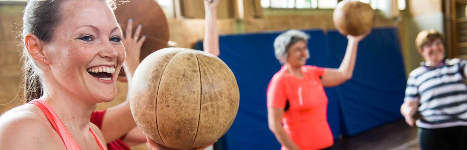 Eine lächelnde Frau in rosa-pinkfarbener Sportbekleidung hält einen braunen Medizinball in ihrer linken Hand hoch. Im Hintergrund sieht man weitere Frauen mit Medizinbällen.