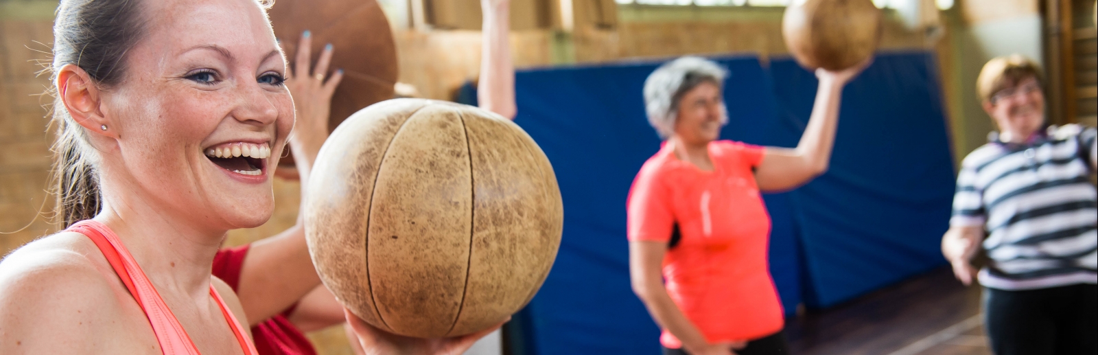 Eine lächelnde Frau in rosa-pinkfarbener Sportbekleidung hält einen braunen Medizinball in ihrer linken Hand hoch. Im Hintergrund sieht man weitere Frauen mit Medizinbällen.