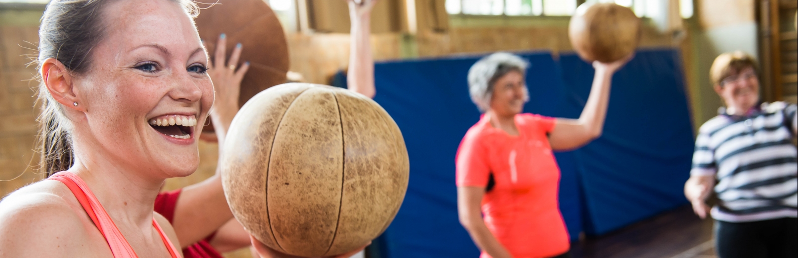 Lachende Frau in einer Turnhalle, sie hält einen Medizinball hoch, mehrere Frauen stehen im Hintergrund.