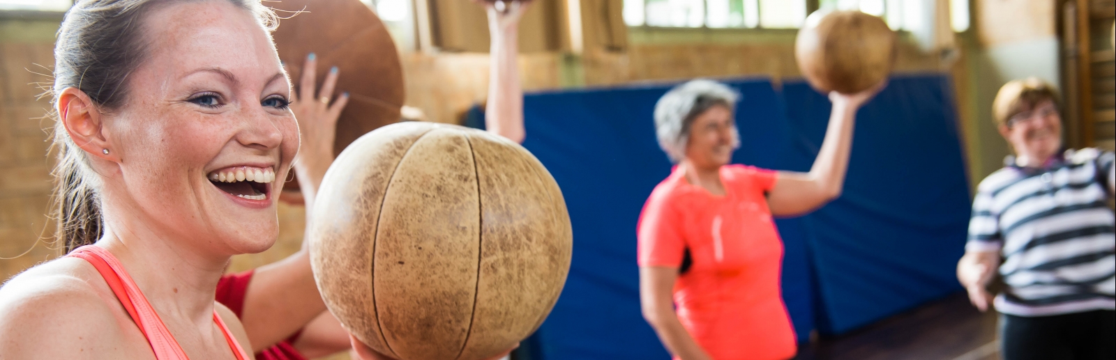 Lachende Frau in einer Turnhalle, sie hält einen Medizinball hoch, mehrere Frauen stehen im Hintergrund.