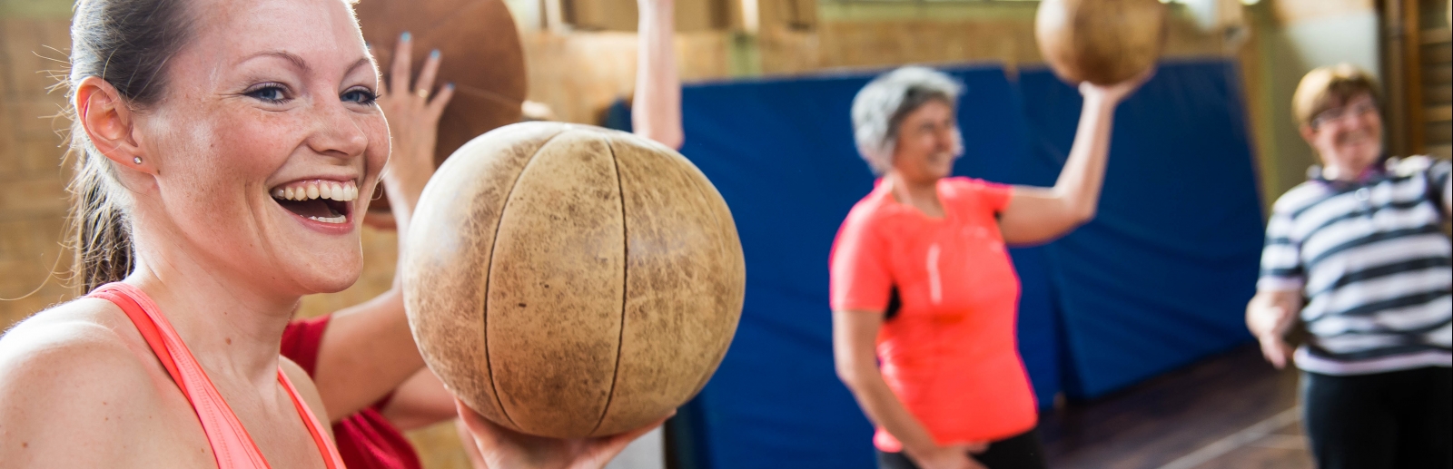 Eine lächelnde Frau in rosa-pinkfarbener Sportbekleidung hält einen braunen Medizinball in ihrer linken Hand hoch. Im Hintergrund sieht man weitere Frauen mit Medizinbällen.