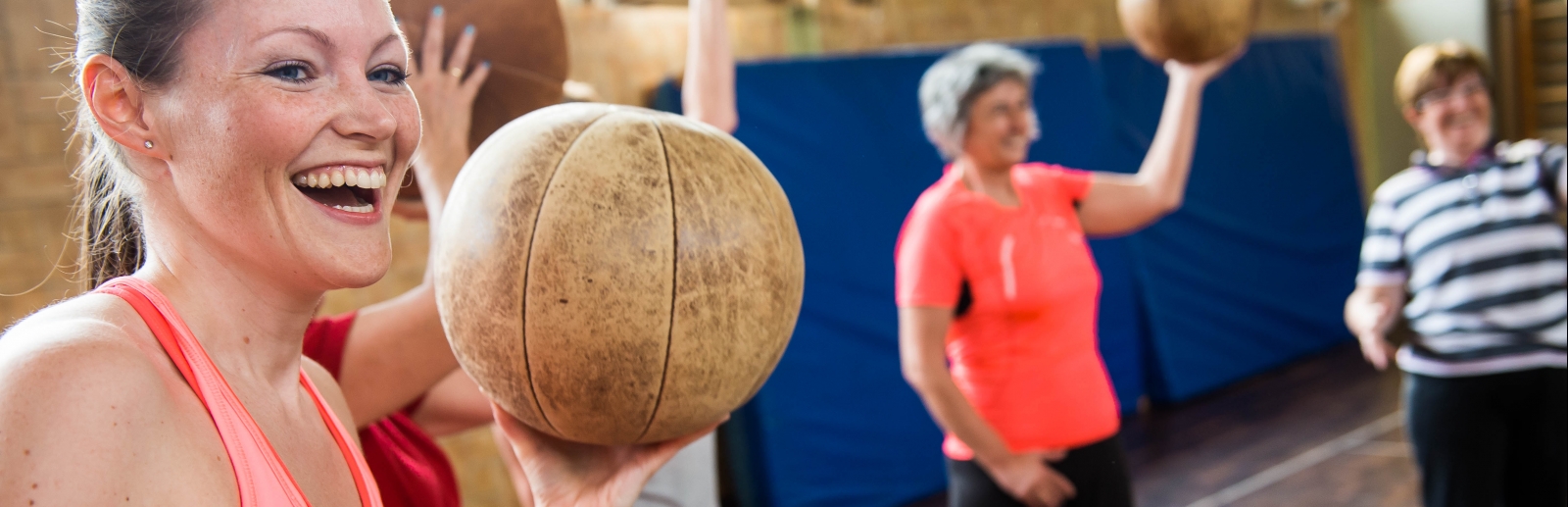 Eine lächelnde Frau in rosa-pinkfarbener Sportbekleidung hält einen braunen Medizinball in ihrer linken Hand hoch. Im Hintergrund sieht man weitere Frauen mit Medizinbällen.