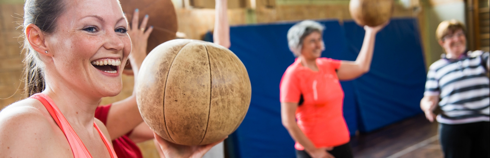 Eine lächelnde Frau in rosa-pinkfarbener Sportbekleidung hält einen braunen Medizinball in ihrer linken Hand hoch. Im Hintergrund sieht man weitere Frauen mit Medizinbällen.