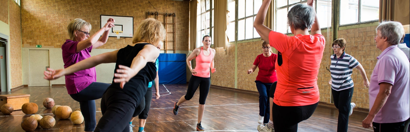 Neun Damen in Sportbekleidung stehen in der Mitter einer Sporthalle im Kreis und machen Aufwärmübungen.