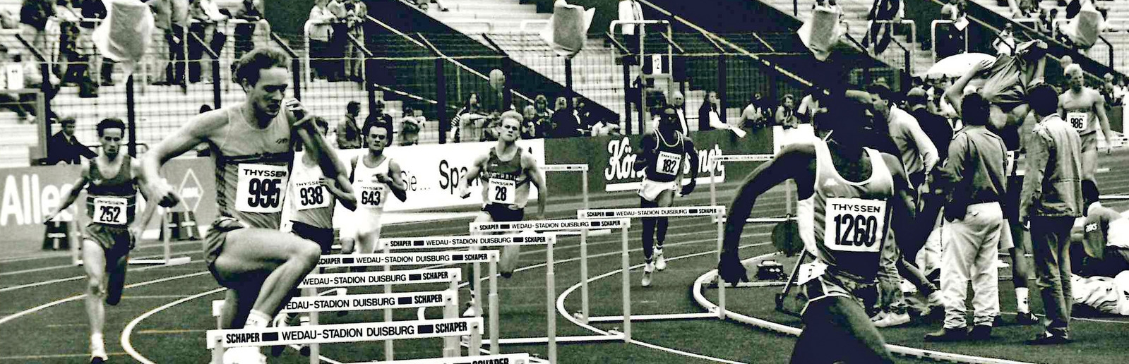 Das schwarz-weiß Foto zeigt mehrere Sportler auf Tartanbahnen beim Hürdenlauf, im Vordergrund springt gerade ein Sportler über eine Hürde