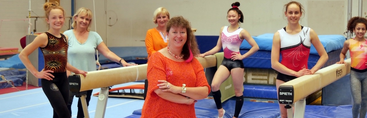 Staatssekretärin Milz in orangefarbenem T-Shirt und Sporthose mit sechs Damen in einer Turnhalle. Zwischen den Damen stehen zwei Schwebebalken, auf dem Boden liegen blaue Bodenmatten.