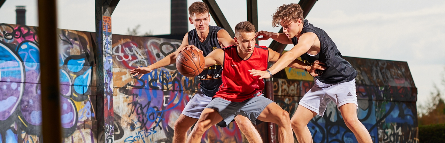 3 Sportler spielen Basketball in eienr Sporthalle, die Wand ist voll verglast.