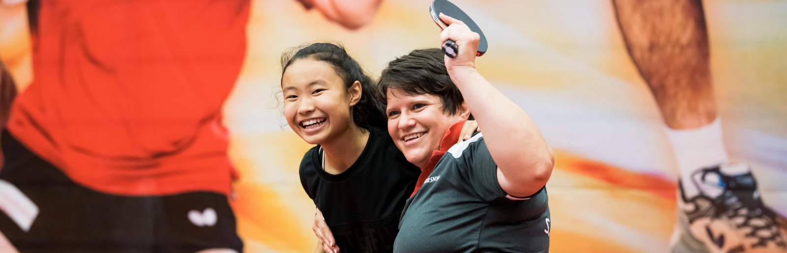 Zwei Tischtennisspielerinnen freuen sich, eine von ihnen sitzt im Rollstuhl. Im Hintergrund an der Wand ein großes Bild von einem Tischtennisspieler im roten T-Shirt und schwarzer, kurzer Hose.