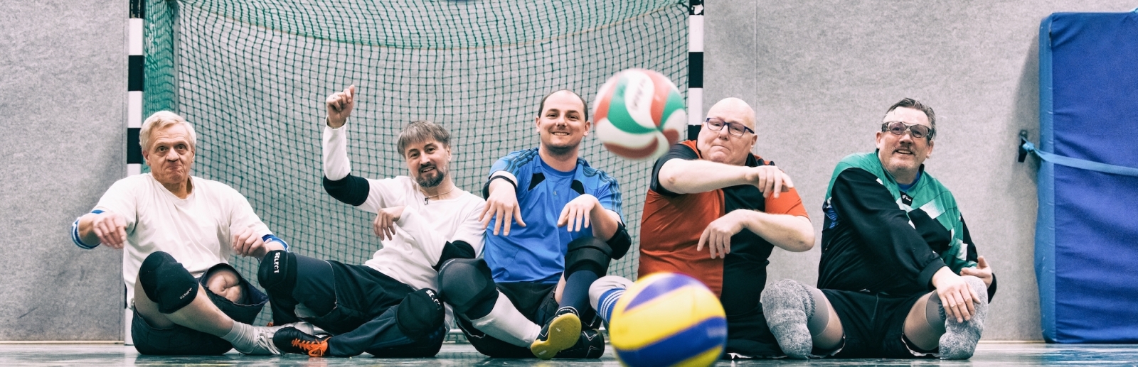 Fünf Sportler sitzend in einer Halle vor einem Tor, werfen Bälle