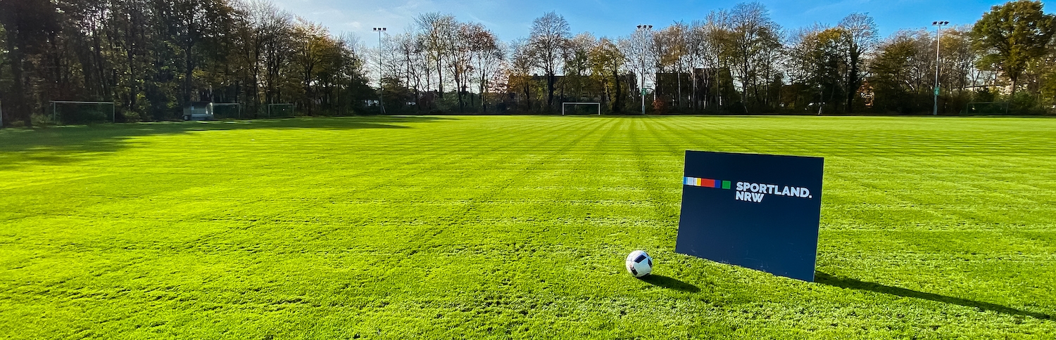 Rasenfläche mit dunkelblauem Sportland.NRW-Schild und einem Fußball 
