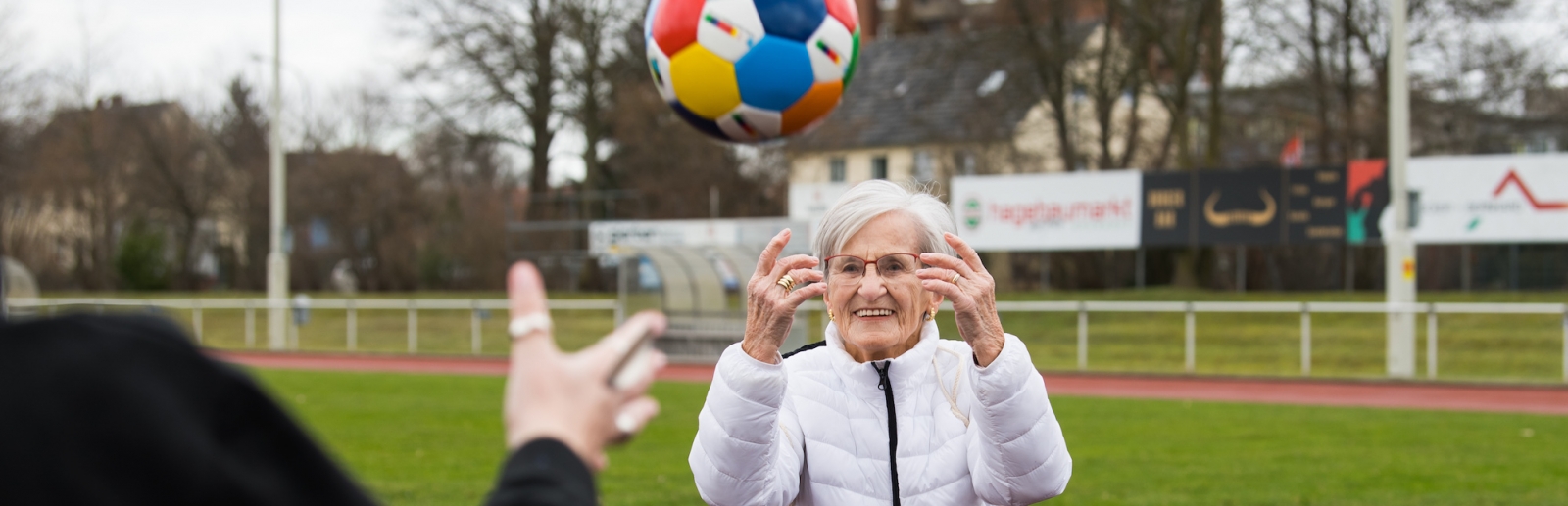 Frau Erika Rischko in weißer Daunenjacke und schwarzer Hose wirft der Staatsekretärin Andrea Milz in schwarzen Sportbekleidung einen bunten Sportland.NRW-Fußball zu. Die sechseckigen Felder des Fußballs haben die Sportland.NRW-Farben Weiß, Rot, Gelb, Grün, Blau und Dunkelblau.