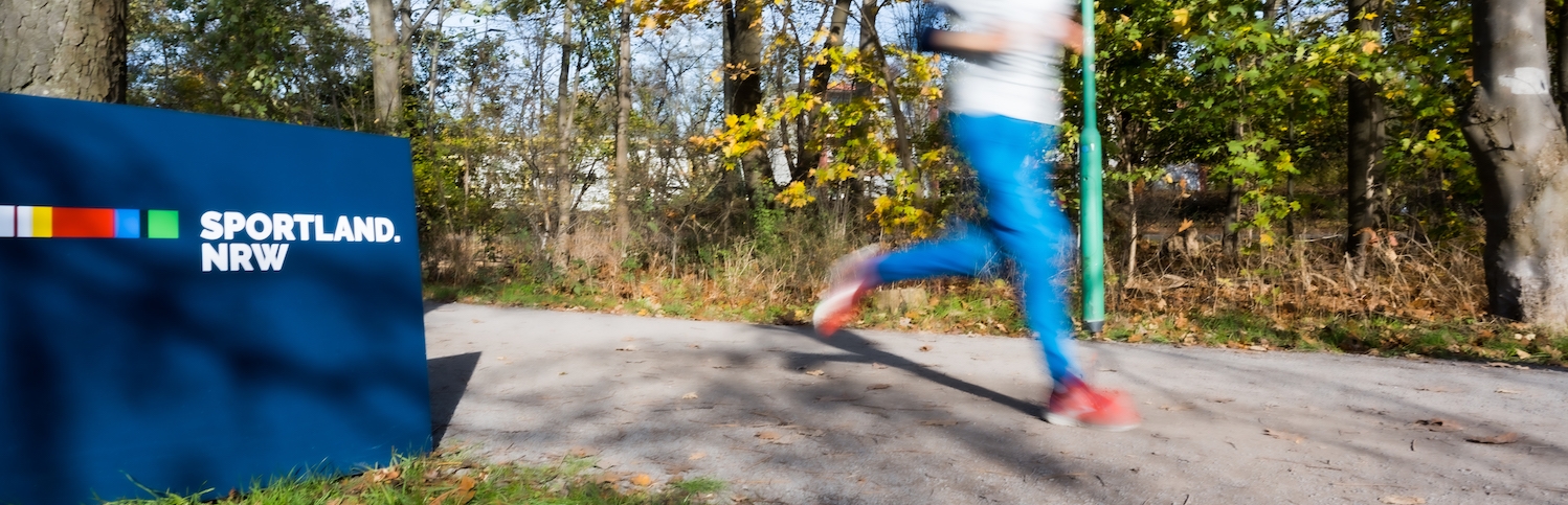 Ein Jogger läuft an einem Sportland.NRW-Schild vorbei