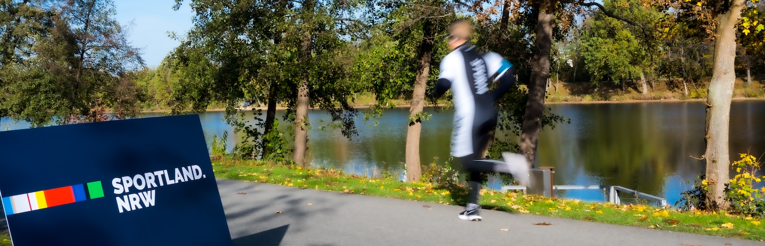 Blaues Sportland.NRW-Schild mit einem vorbeilaufenden Jogger