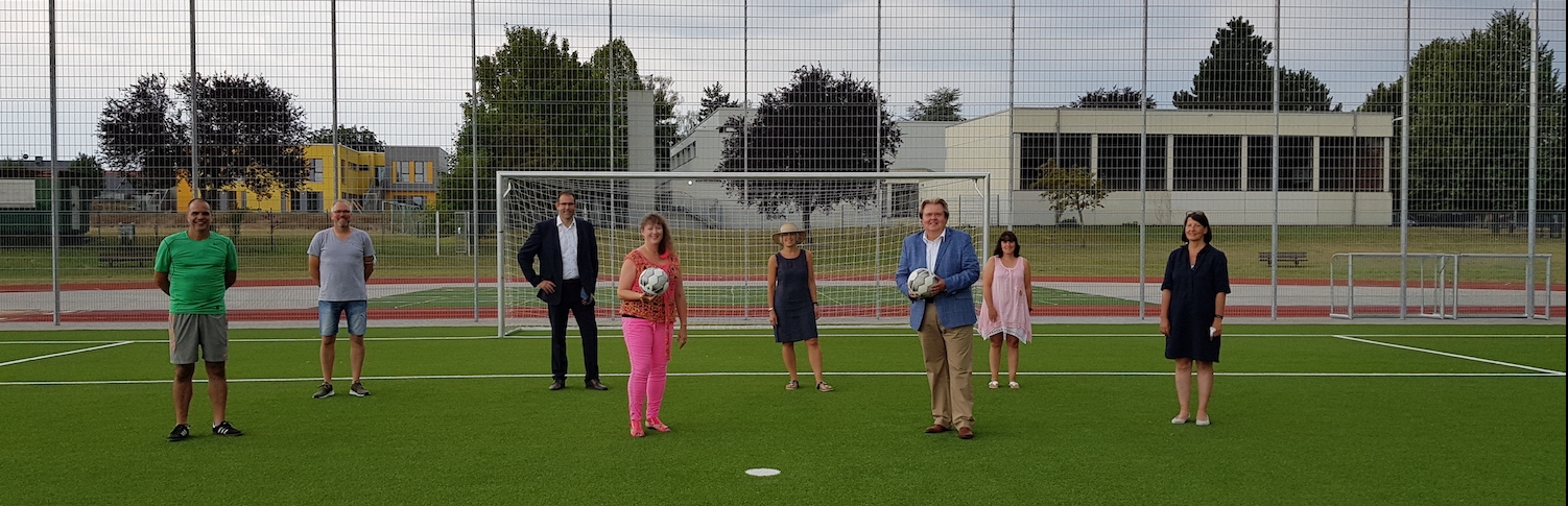 Staatssekretärin Andrea Milz mit Vereinsmitgliedern auf einen Fußball-Rasenplatz. Hinter ihnen steht ein weißes Fußballtor.