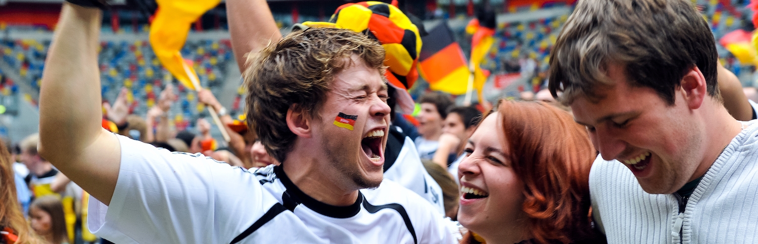 Jubelnde Fans mit Deutschland-Flaggen i einem Stadion. Im Vordergrund ein Fan in weiß-schwarzem Trikot und Deutschland-Flagge auf der Wange.