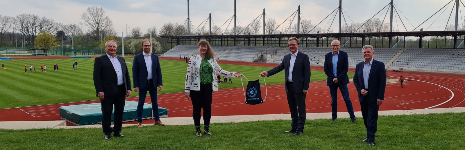 Staatssekretärin Andrea Milz steht mit 5 weiteren Menschen vor einer Sportstätte mit Tartanbahnen und einem Rasenplatz in der Mitte.