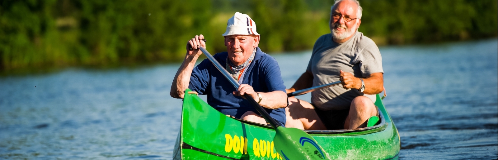 Zwei Senioren paddeln gemeinsa in einem grünen Kanu über ein ruhiges Gewässer.