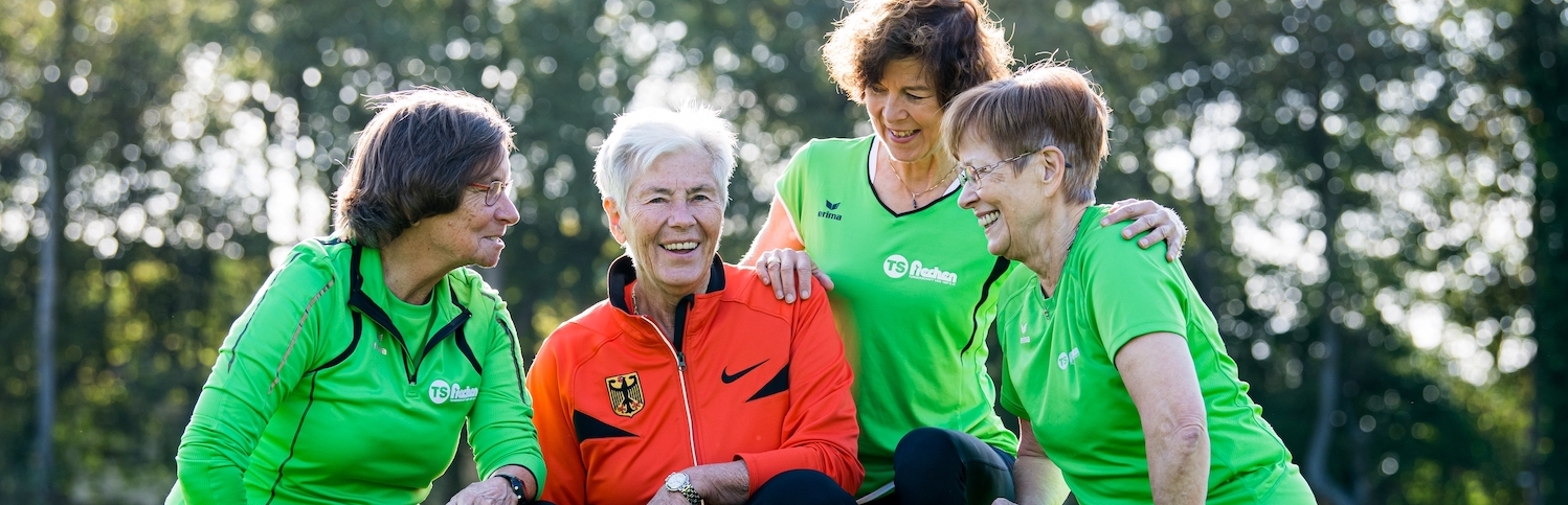 Vier ältere Damen bei einer Teambesprechung beim Sport. Drei Damen haben ein grünes Oberteil an, eine Dame in der Mitte hat ein rotes Oberteil an. Alle haben schwarze Sporthosen an.