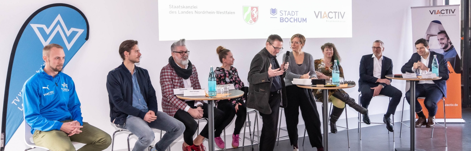 Staatssekretärin Andrea Milz sitzt mit Teilnehmern der Pressekonferenz auf Hockern an Rundtischen. Hinter ihnen eine weiße Rückwand mit einer Präsentation.