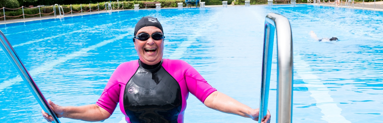 Eine Schwimmerin im pink-schwarzen Neoprenanzug, mit schwarzer Badekappe und schwarzer Schwimmbrille steigt aus einem offenen Schwimmbecken, ihre beiden Hände jeweils links und rechts am Geländer.