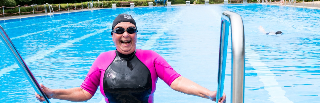 Eine Schwimmerin im pink-schwarzen Neoprenanzug, mit schwarzer Badekappe und schwarzer Schwimmbrille steigt aus einem offenen Schwimmbecken, ihre beiden Hände jeweils links und rechts am Geländer.