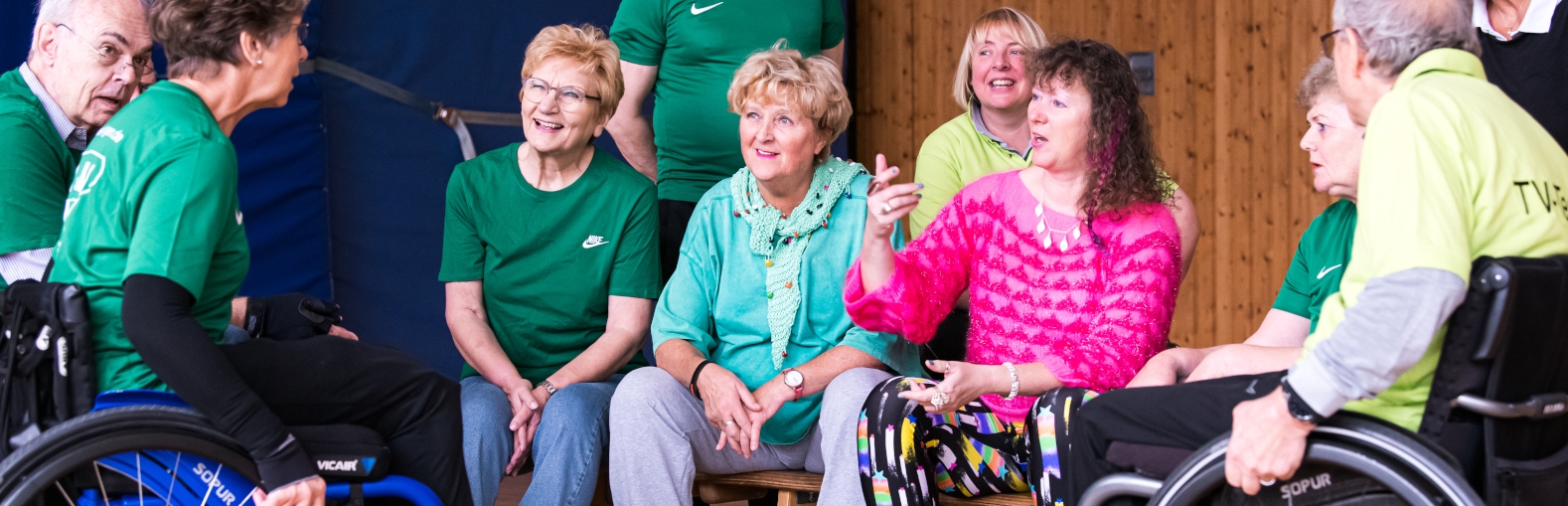 Staatssekretärin Milz sitzt mit mehreren Sportlern in einer Sporthalle. Links sitzt eine Dame im Rollstuhl, rechts ein Herr im Rollstuhl.