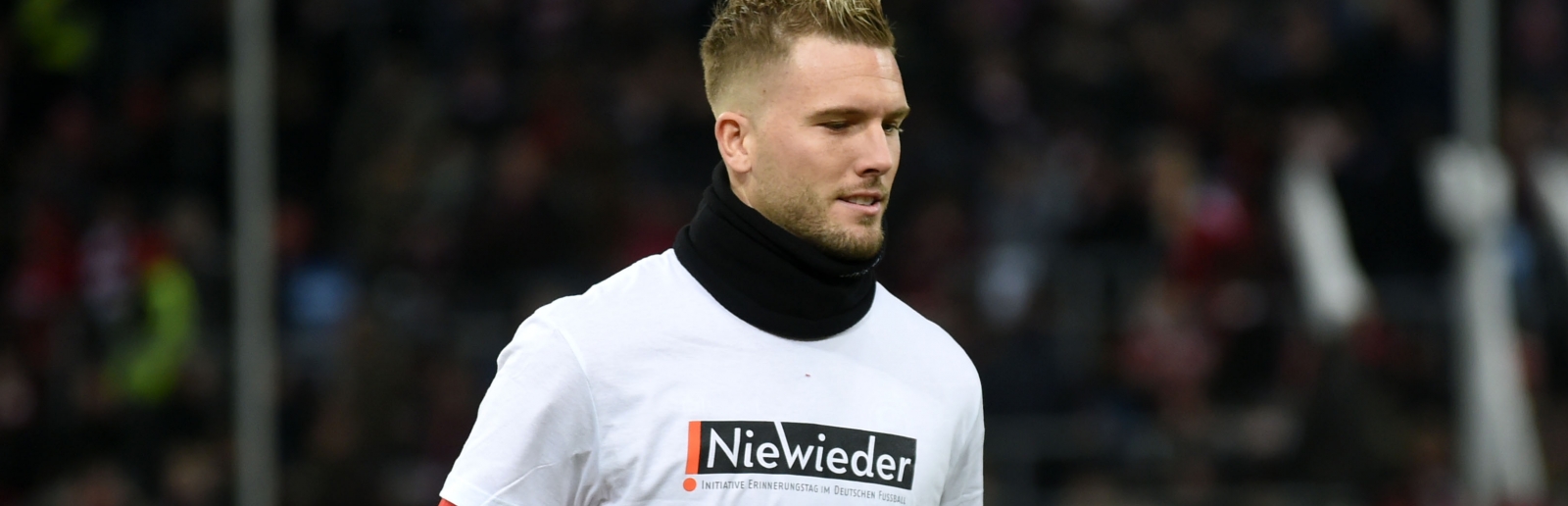Ein Fußballer mit weiß-rotem FC-Trikot und schwarzer Hose geht über den Sportplatz, auf seinem T-Shirt steht Weiß auf Schwarz "Nie Wieder".