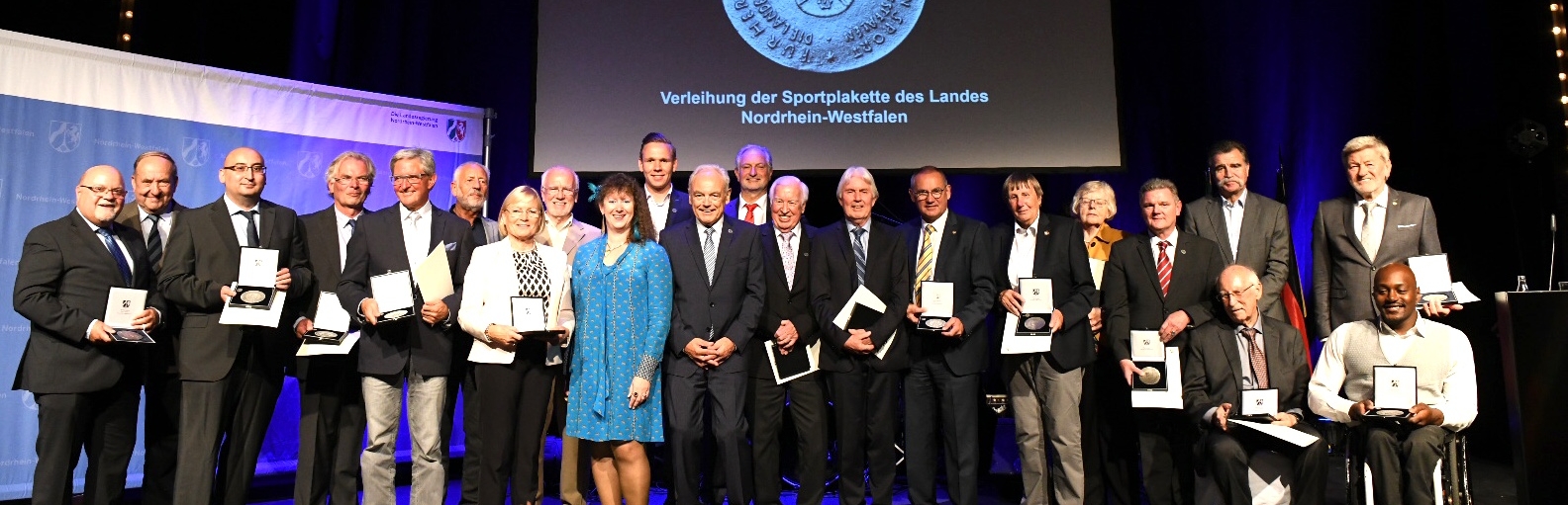 Gruppenfoto auf einer Bühne mit Staatssekretärin Milz im hellblauen Kleid und allen Trägern der Sportplakette.