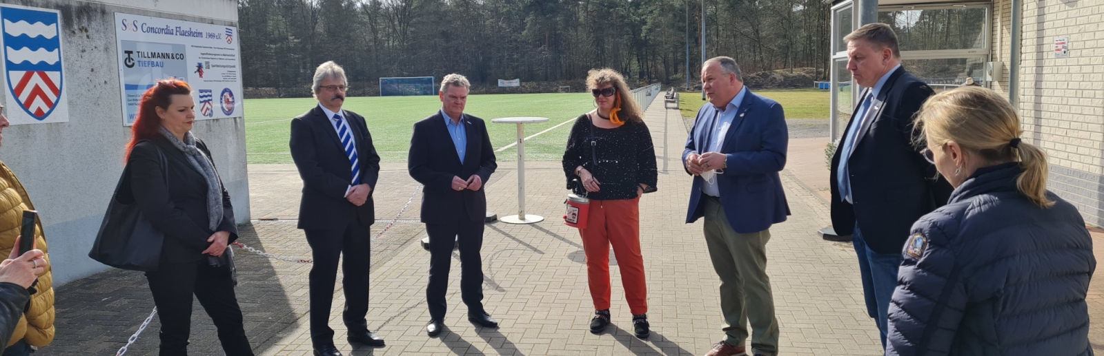 Staatssekretärin Milz steht mit mehreren Personen im Halbkreis vor einem Rasensportplatz.