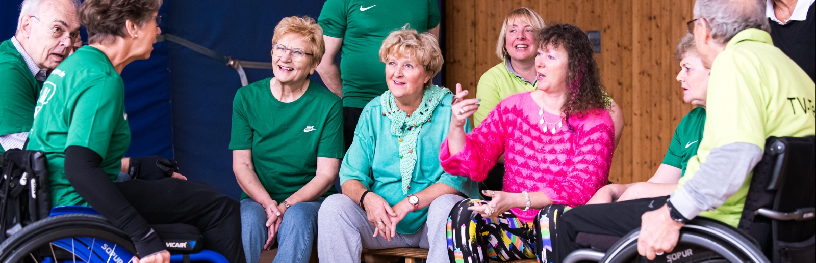 Staatssekretärin Milz im Gespräch mit Sportlern auf einer Sportbank in einer Sporthalle sitzend. Eine Sportlerin und ein Sportler sitzen in Rollsüthlen.