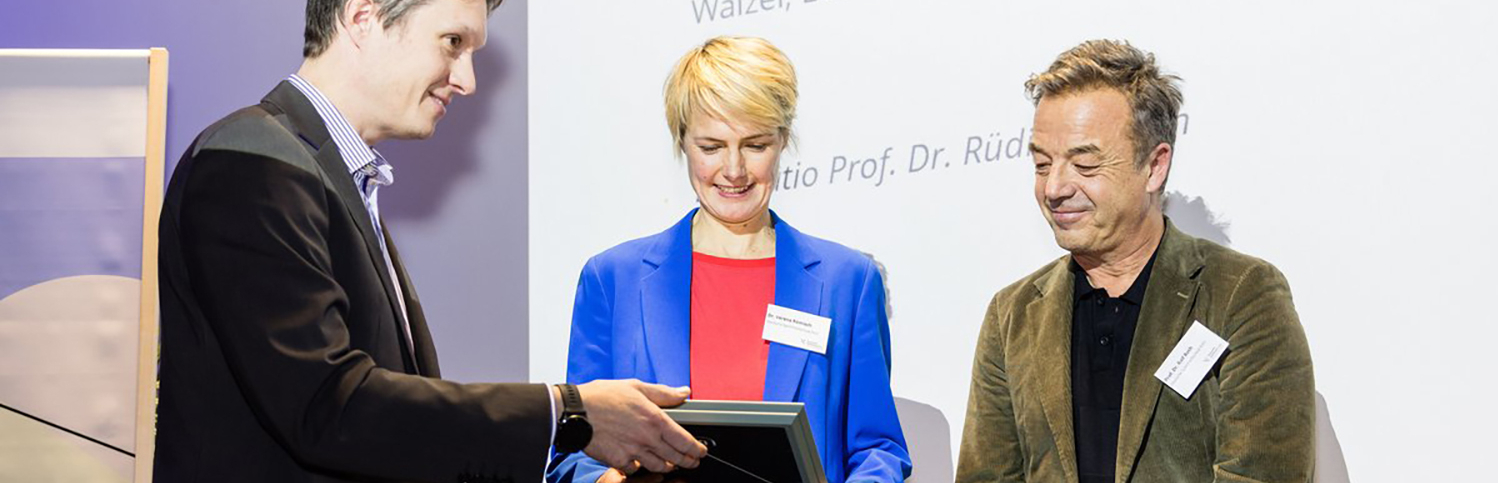 Drei Personen, in der Mitte eine Frau, bei der Übergabe einer Urkunde