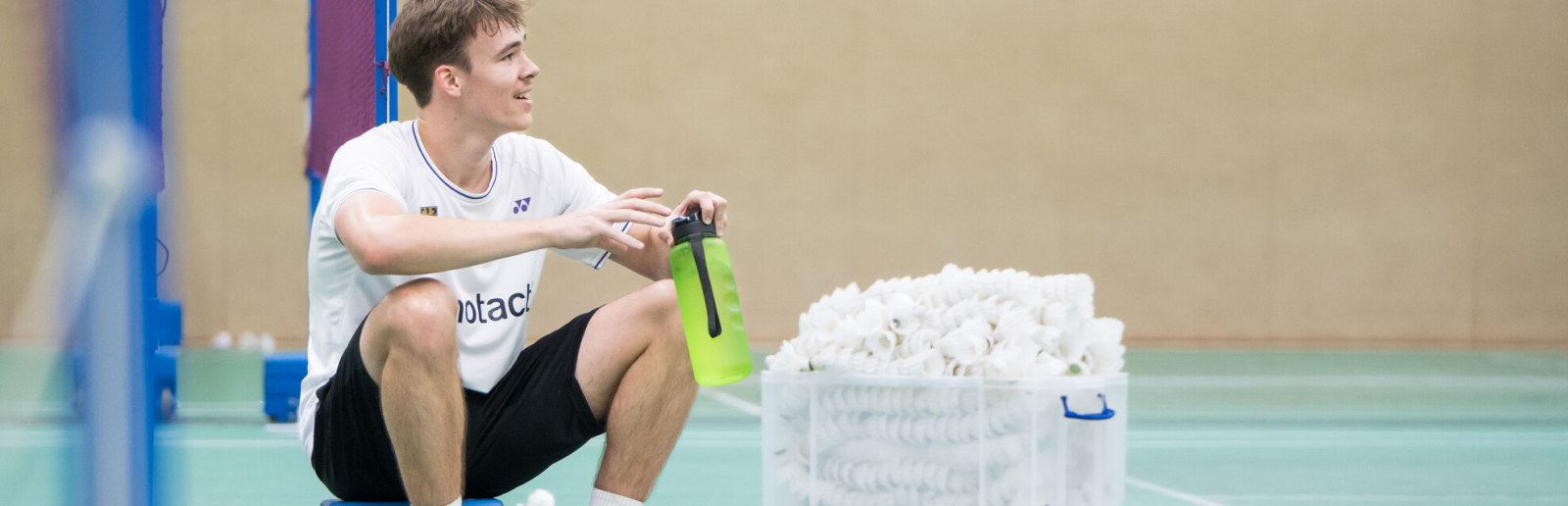 Linus sitzt mit angezogenen Knien in der Badmintonhalle am Netz