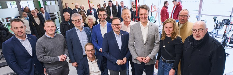 Hendrick Wüst inmitten mehrerer Personen, davon einer im Rollstuhl, in einem Raum mit Fenstern im Hintergrund