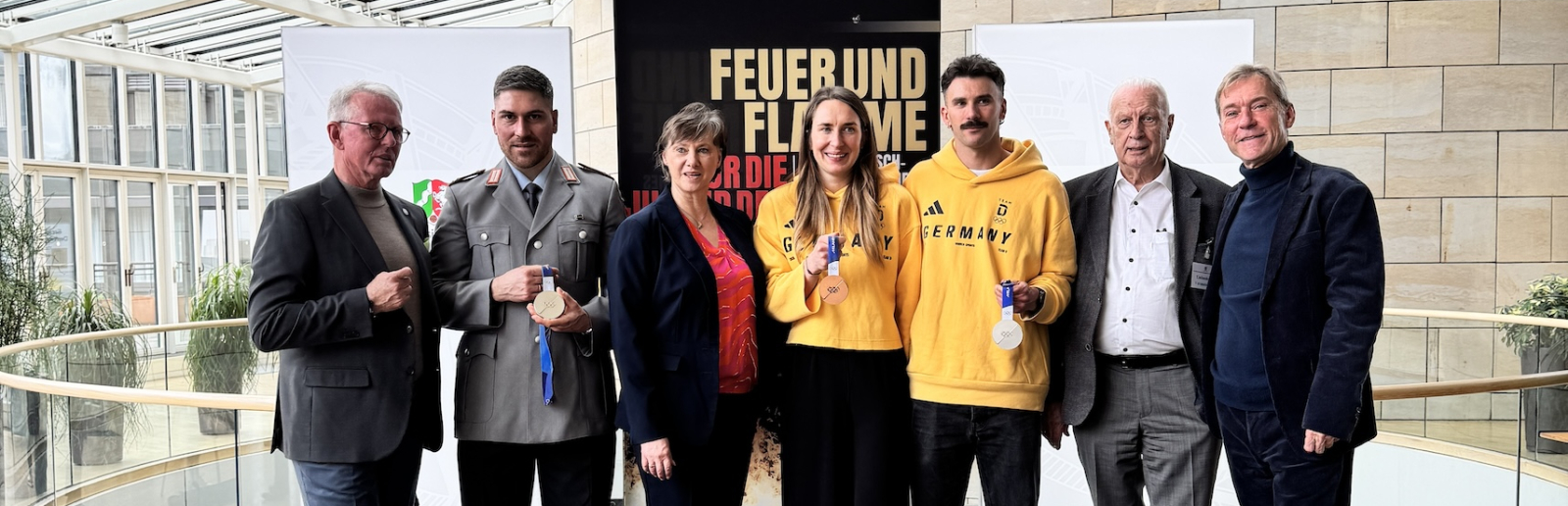 Sieben Personen auf einem schwarzen Podest im Landtag NRW mit vielen Fenstern im Hintergrund