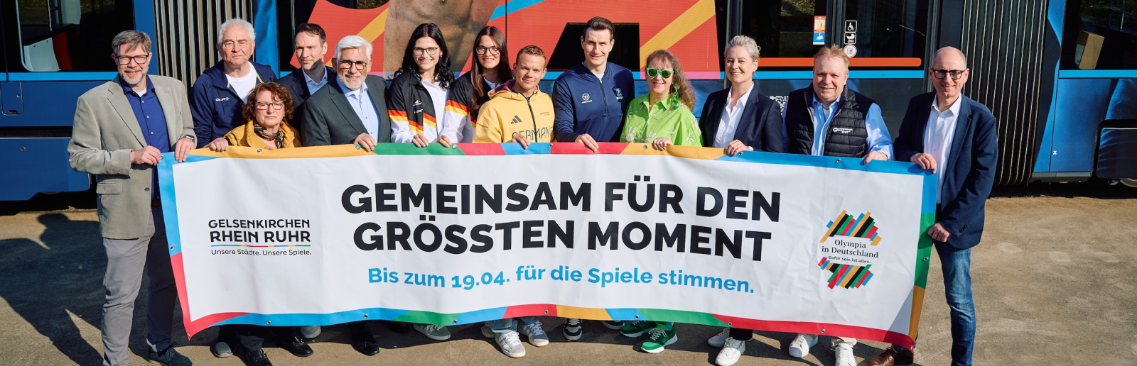 Andrea Milz und weitere Personen mit einem Banner der KRR-Bewerbung vor einer Straßenbahn im "Olympia-Look"