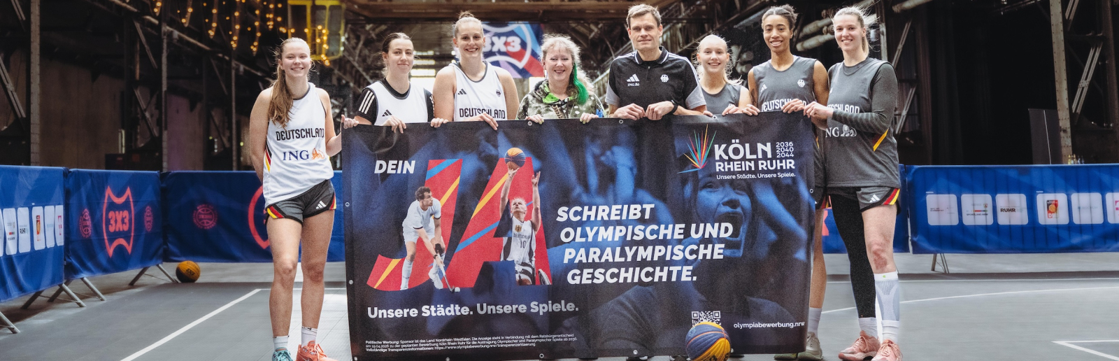 Andrea Milz mit 3x3-Aktiven und weiteren Personen in einer Sporhalle, sie halten ein Banner für die Bewerbung KölnRheinRuhr