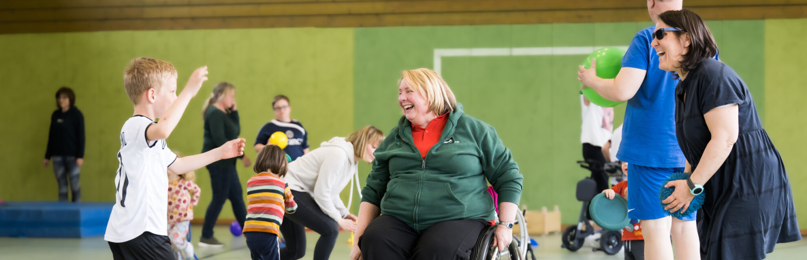 Ältere Frau in Sportkleidung im Rollstuhl ist umgeben von jüngeren Sportlern in eienr Sporthalle