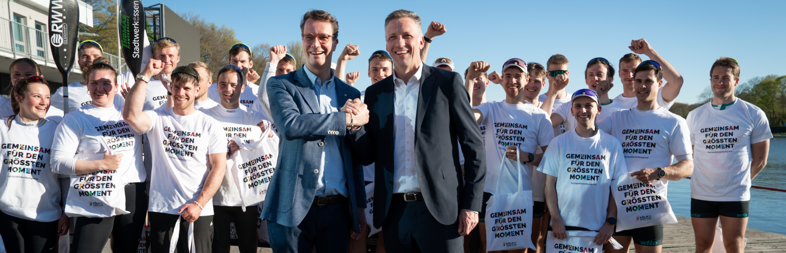 Hendrik Wüst umgeben von Ruderen in weißen Shirts mit der Aufschrift "Gemeinsam für den Größten Moment"