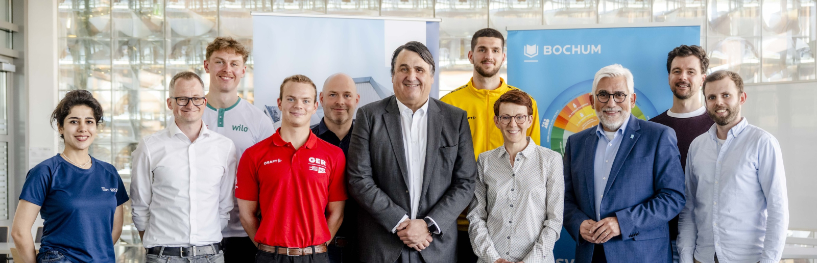 Elf Personen in einem Halbkreis vor zwei Bannern, hinter ihnen eine Trennwand aus Glasbausteinen