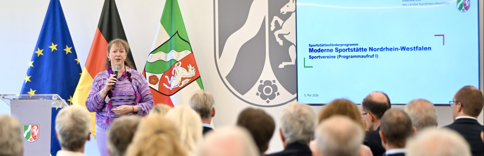Andrea Milz vor drei Fahnen und Videobildschirm, sie spricht zu Teilnehmenden einer Pressekonferenz