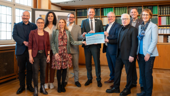 Gruppe von Personen in einem Raum mit Holzboden, ein Mann in der Mitte hält einen Bescheid in der Hand